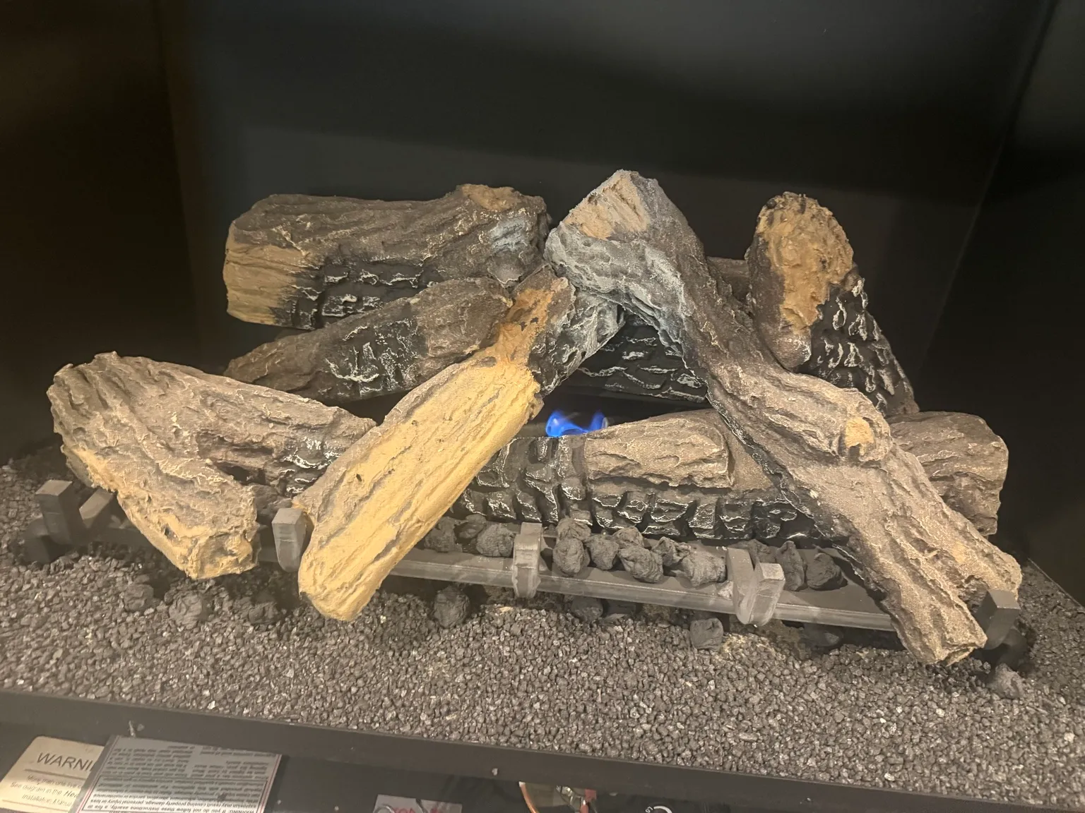 Gas log set with a blue burner flame, on a bed of gravel inside a dark firebox.