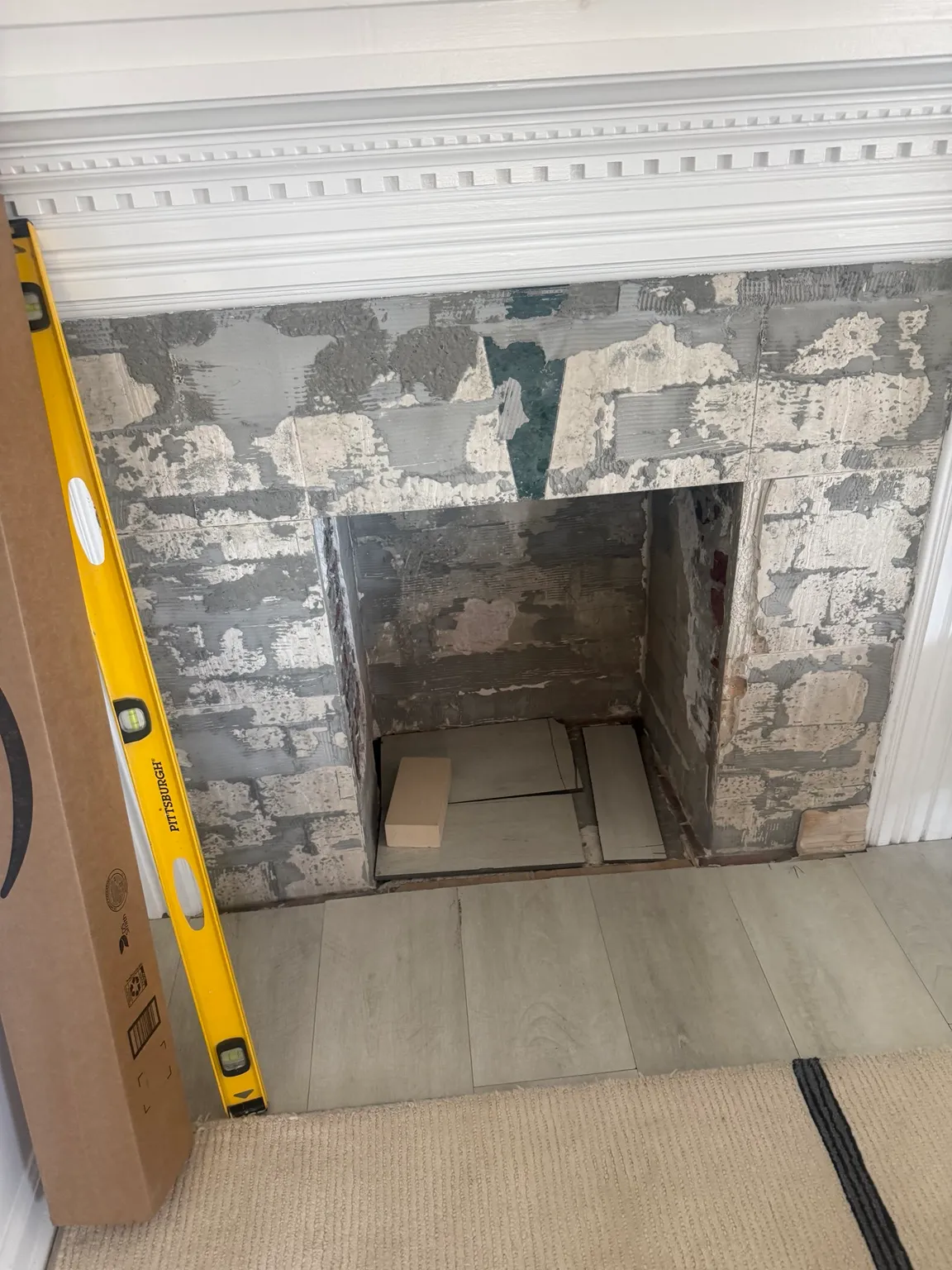 Fireplace opening with its surround stripped back to patchy grey substrate, a yellow level standing at one side and sheets of cardboard laid inside.