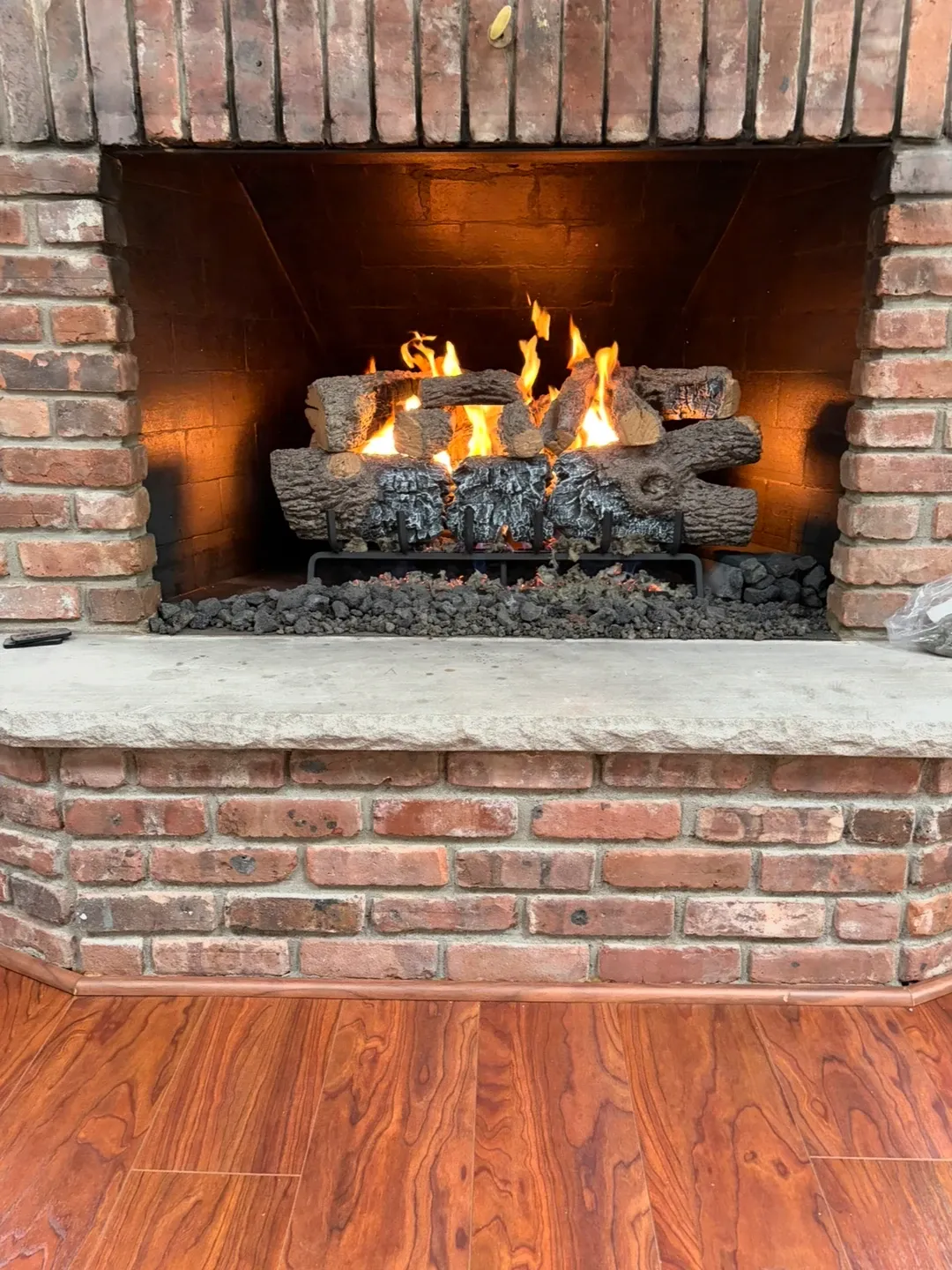 Gas log set alight in a red brick fireplace, flames over a ceramic log set on a bed of lava rock above a concrete hearth slab.